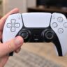 Hand holding a DualSense wireless controller in a living room.