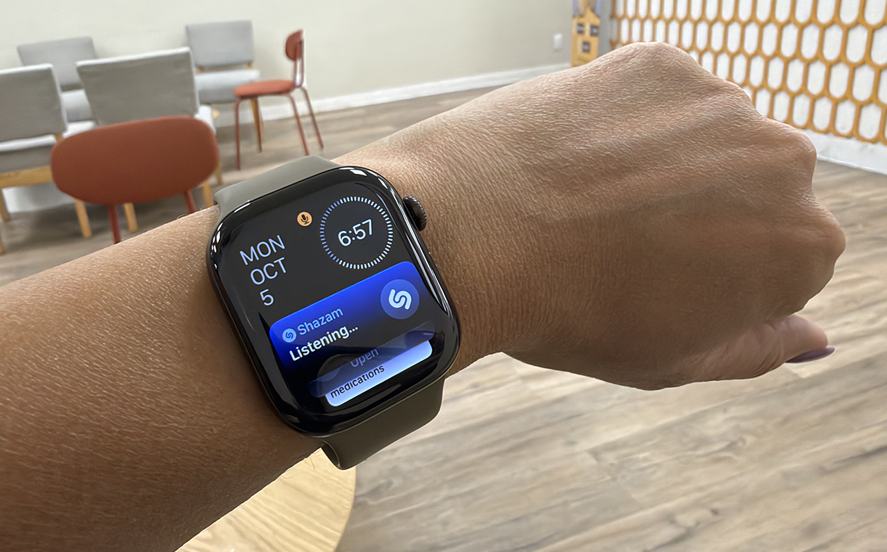 An arm showing an Apple Watch Series 12 in a cafe with Shazam listening for the tune.